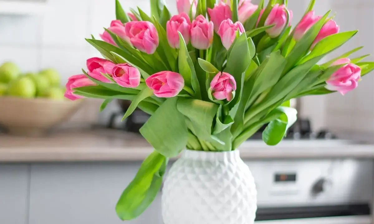 Różowe tulipany w białym wazonie, idealne na stół weselny. W tle miska z zielonymi jabłkami i kuchenny blat.