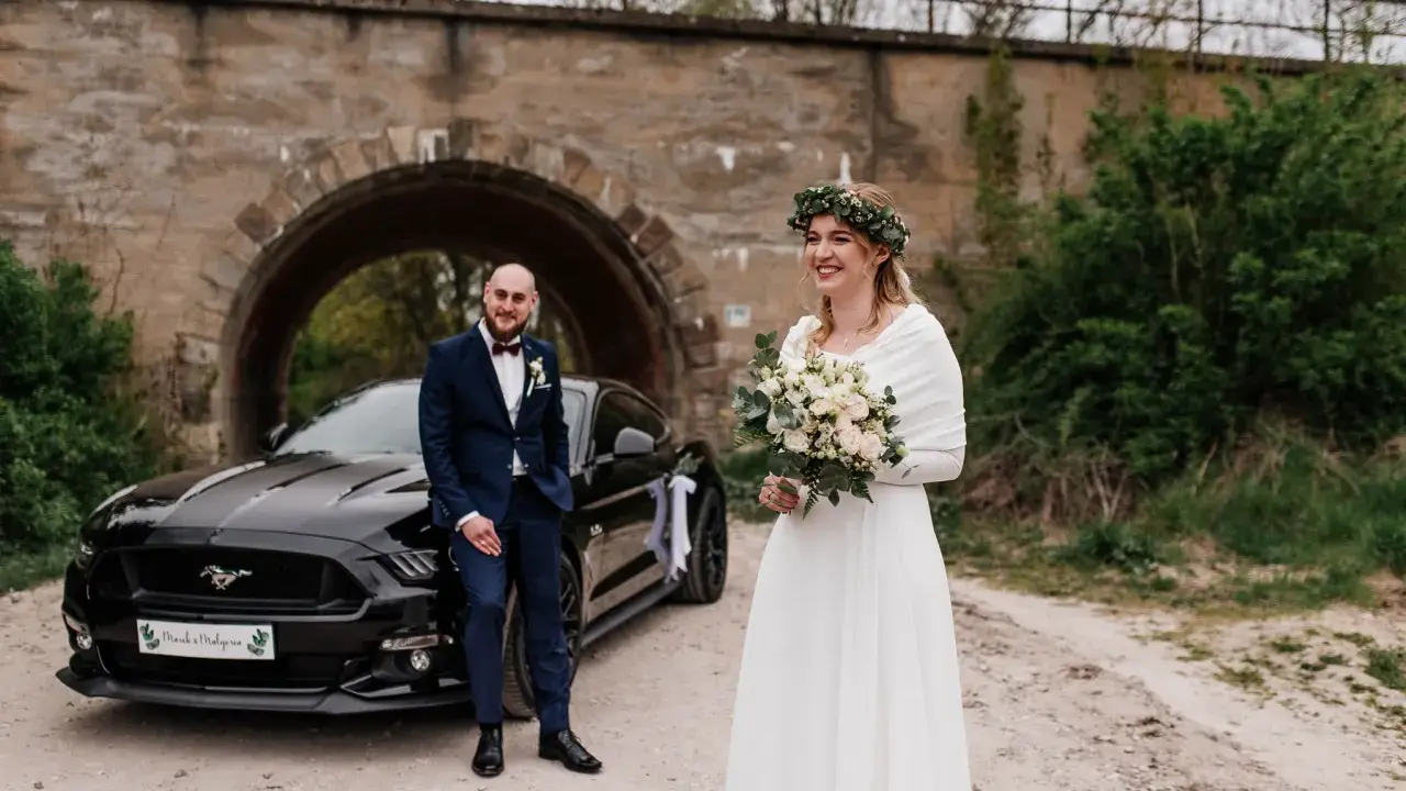 Para nietypowe sesje ślubne pozuje przy czarnym Mustangu pod kamiennym wiaduktem. Panna młoda w białej sukni i wianku z zieleni, pan młody w granatowym garniturze.