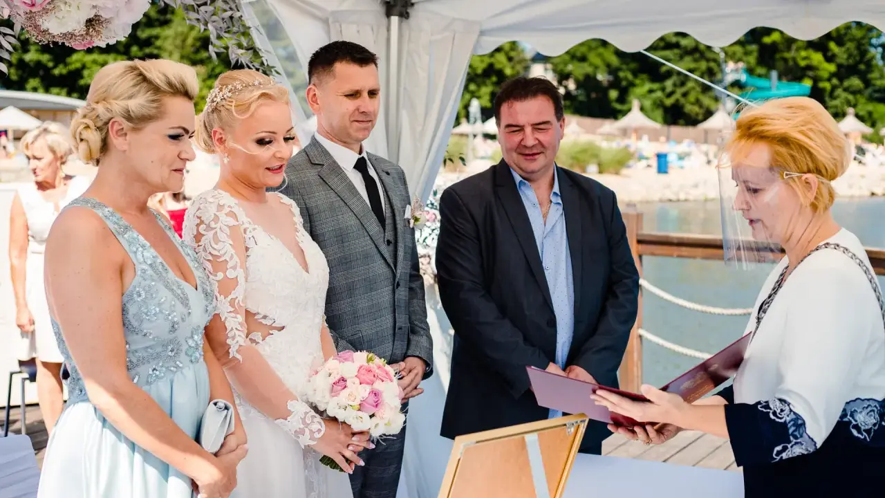 Urzędniczka w przyłbicy wręcza parze młodej dokumenty, symbolizujące ich licencję na ślub.