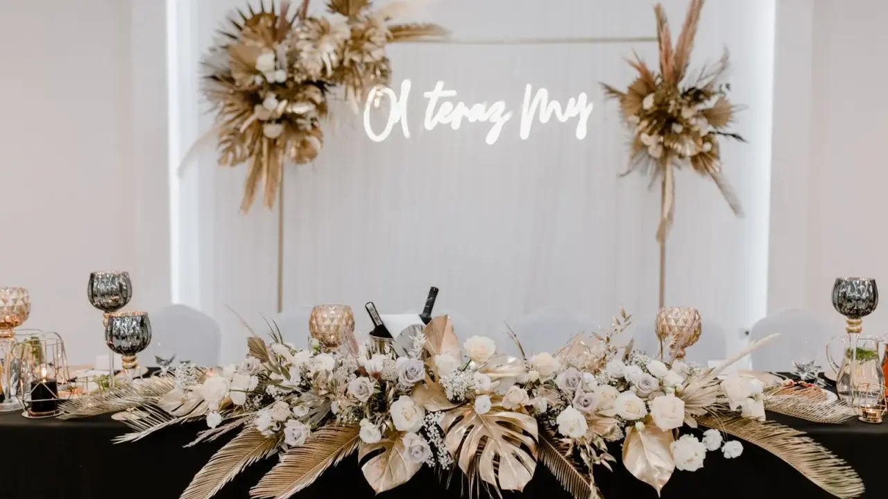 Elegancka ścianka za młodymi boho z napisem "O, teraz My". Dekoracja stołu z bukietami kwiatów, złotymi liśćmi i świecami.
