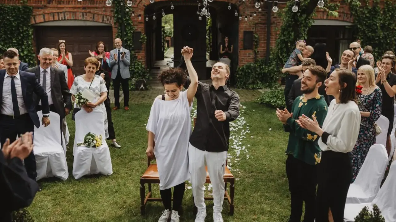 Para młoda świętuje ślub cywilny na luzie, trzymając się za ręce. Goście klaszczą, ciesząc się radosnym momentem.