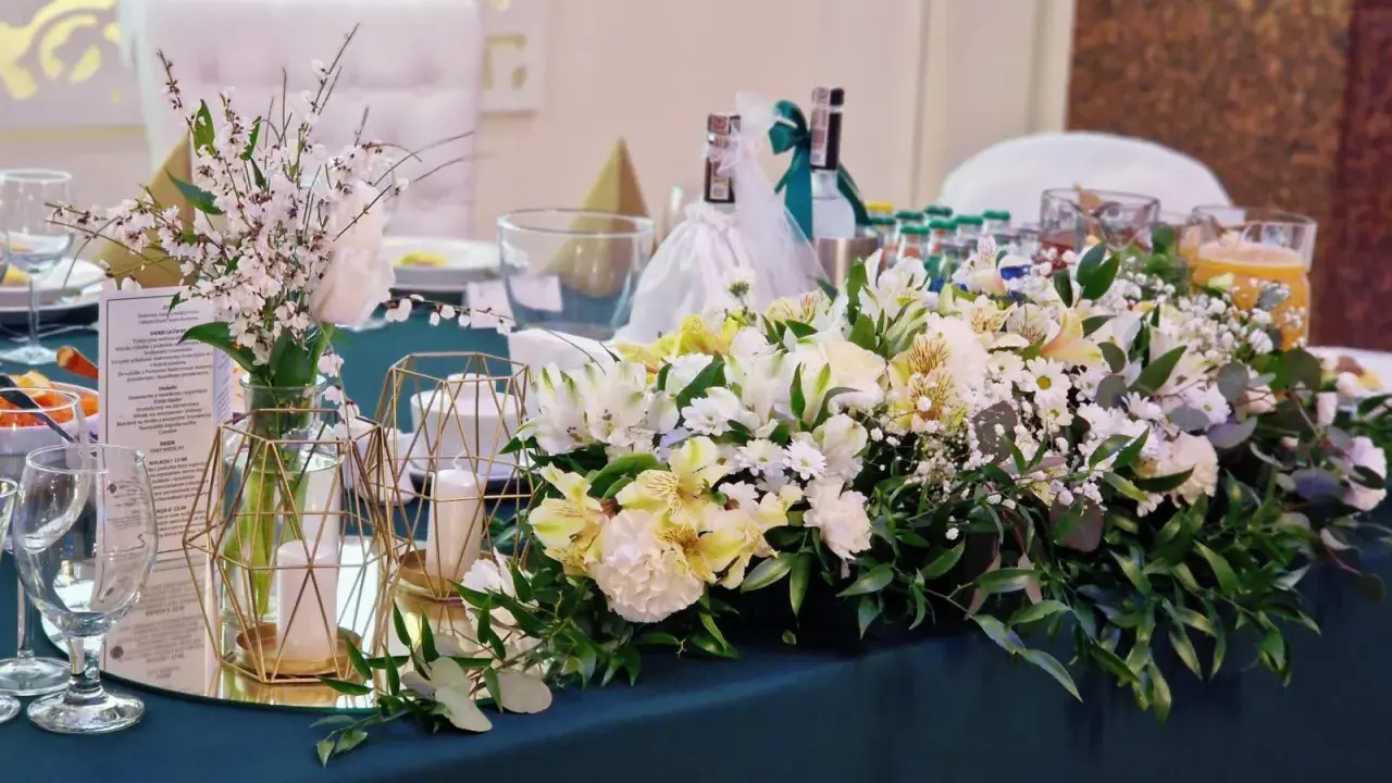 Elegancki pomysł na wesele: stół udekorowany bukietami białych i żółtych kwiatów, świecami w złotych lampionach i butelkami z napojami.