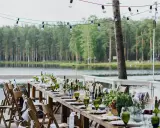 Ustawiony długi stół z zielonymi kieliszkami i kwiatami nad jeziorem. Girlanda żarówek tworzy magiczną atmosferę, idealną na tanie wesele w plenerze.