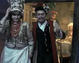 Młoda para w strojach ludowych, o czym jest wesele. W tle widać gości przy stole.