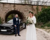 Para nietypowe sesje ślubne pozuje przy czarnym Mustangu pod kamiennym wiaduktem. Panna młoda w białej sukni i wianku z zieleni, pan młody w granatowym garniturze.