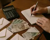 Rachunki, koperty i polskie stuzłotówki na stole. Czy pieniądze z wesela można wpłacić na konto? Dłonie zapisują wydatki.