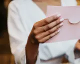 Dłoń z pierścionkiem trzyma różową kopertę. Czy to zaproszenie na wesele?