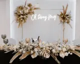 Elegancka ścianka za młodymi boho z napisem "O, teraz My". Dekoracja stołu z bukietami kwiatów, złotymi liśćmi i świecami.
