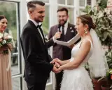 Para młoda trzyma się za ręce podczas ceremonii. W tle widać świadkową z bukietem i urzędnika. To efekt pracy świetnej organizatorki wesel.