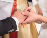 Dłoń panny młodej zakłada obrączkę na palec pana młodego. W tle widać księdza w szacie liturgicznej. To piękny ślub w innej parafii.