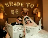 Para młoda czyta gazetę "The Wedding Post" z nagłówkami o ślubie. Czyżby to zapowiedź, ile trwają poprawiny?