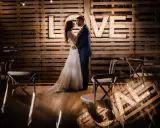 Para młoda w objęciach na tle napisu "LOVE" z żarówek. Piękny plener ślubny z drewnianą ścianą i krzesłami.