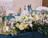 Elegancki pomysł na wesele: stół udekorowany bukietami białych i żółtych kwiatów, świecami w złotych lampionach i butelkami z napojami.