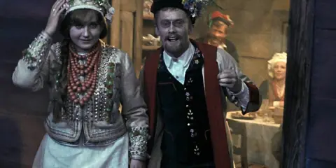Młoda para w strojach ludowych, o czym jest wesele. W tle widać gości przy stole.