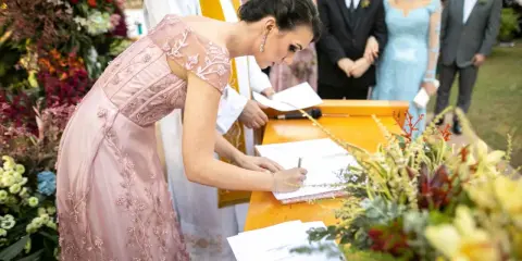Świadek na ślubie cywilnym podpisuje dokumenty. Kobieta w różowej sukni z haftami pochyla się nad stołem, by złożyć swój podpis.