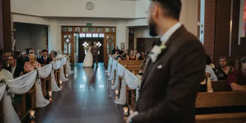 Panna młoda z ojcem idzie do ołtarza. W kościele panują uroczyste warunki ślubu kościelnego, z gośćmi siedzącymi w ławkach.