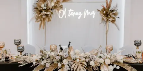 Elegancka ścianka za młodymi boho z napisem "O, teraz My". Dekoracja stołu z bukietami kwiatów, złotymi liśćmi i świecami.