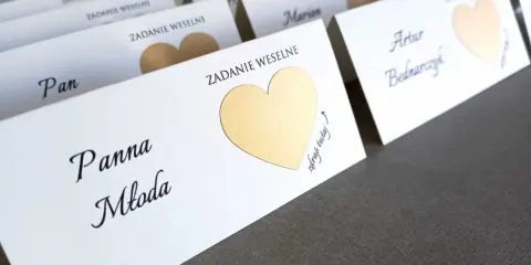 Białe karty z imionami gości i złotymi serduszkami kryją śmieszne zadania dla gości weselnych.