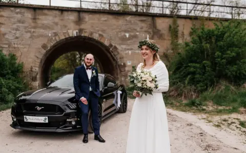 Para nietypowe sesje ślubne pozuje przy czarnym Mustangu pod kamiennym wiaduktem. Panna młoda w białej sukni i wianku z zieleni, pan młody w granatowym garniturze.