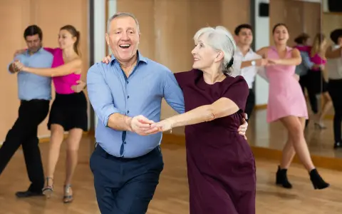 Uśmiechnięta para seniorów uczy się tańczyć w parze. W tle inne pary ćwiczą kroki.