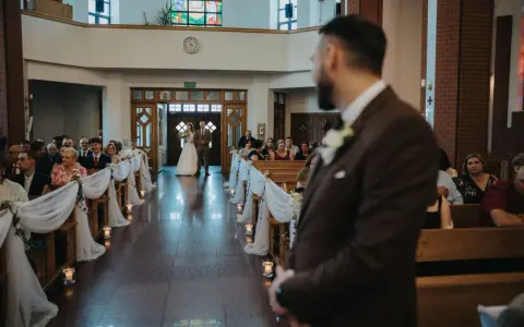 Panna młoda z ojcem idzie do ołtarza. W kościele panują uroczyste warunki ślubu kościelnego, z gośćmi siedzącymi w ławkach.