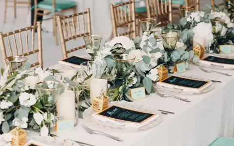 Elegancko nakryty stół z menu i kwiatami. Złote krzesła i zielone kieliszki tworzą atmosferę uroczystego obiadu weselnego zamiast wesela.