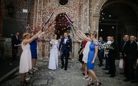 Szczęśliwa para młoda wychodzi z kościoła, obsypana płatkami. Goście z radością świętują wyjście z kościoła ślub.