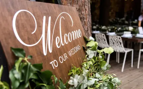 Drewniana tablica "Welcome to Our Wedding" ozdobiona zielenią. W tle widać przygotowane stoły i krzesła, a także obsługę. Opłata ZAIKS za muzykę na weselu.