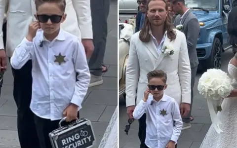 Chłopiec w okularach przeciwsłonecznych, z odznaką i walizką "Ring Security", niesie obrączki do ślubu.