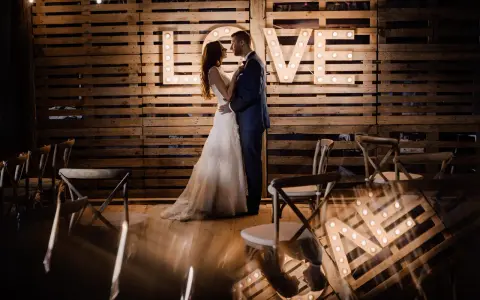 Para młoda w objęciach na tle napisu "LOVE" z żarówek. Piękny plener ślubny z drewnianą ścianą i krzesłami.