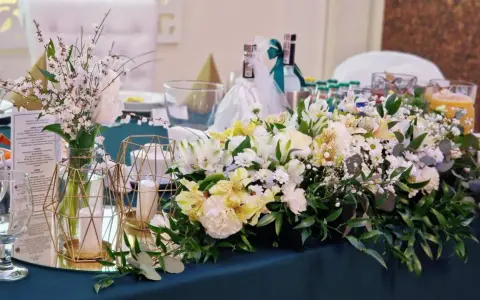 Elegancki pomysł na wesele: stół udekorowany bukietami białych i żółtych kwiatów, świecami w złotych lampionach i butelkami z napojami.