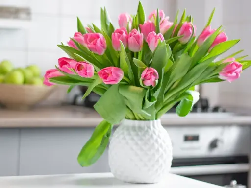 Różowe tulipany w białym wazonie, idealne na stół weselny. W tle miska z zielonymi jabłkami i kuchenny blat.