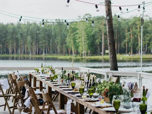 Ustawiony długi stół z zielonymi kieliszkami i kwiatami nad jeziorem. Girlanda żarówek tworzy magiczną atmosferę, idealną na tanie wesele w plenerze.