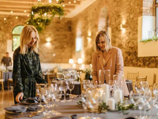 Dwie kobiety przygotowują salę na przyjęcie. Stół nakryty jest eleganckimi naczyniami, tworząc idealny scenariusz wesela.