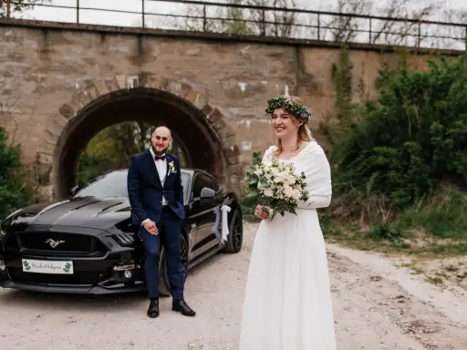 Para nietypowe sesje ślubne pozuje przy czarnym Mustangu pod kamiennym wiaduktem. Panna młoda w białej sukni i wianku z zieleni, pan młody w granatowym garniturze.