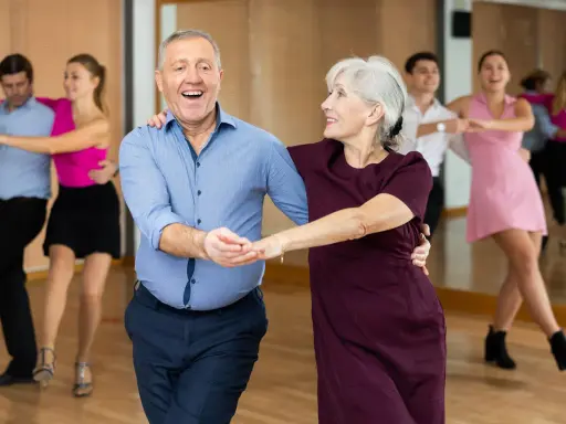 Uśmiechnięta para seniorów uczy się tańczyć w parze. W tle inne pary ćwiczą kroki.