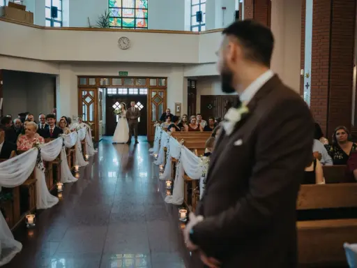 Panna młoda z ojcem idzie do ołtarza. W kościele panują uroczyste warunki ślubu kościelnego, z gośćmi siedzącymi w ławkach.