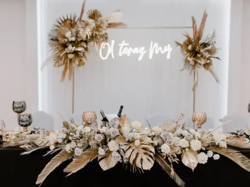 Elegancka ścianka za młodymi boho z napisem "O, teraz My". Dekoracja stołu z bukietami kwiatów, złotymi liśćmi i świecami.