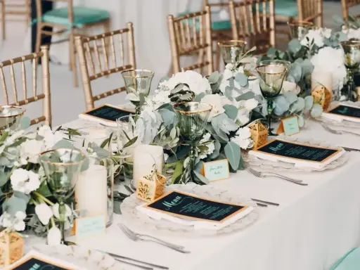 Elegancko nakryty stół z menu i kwiatami. Złote krzesła i zielone kieliszki tworzą atmosferę uroczystego obiadu weselnego zamiast wesela.