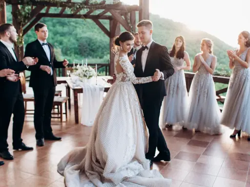 Para Młoda tańczy swój pierwszy taniec. Panna Młoda w pięknej sukni, Pan Młody w eleganckim garniturze. Druhny i świadkowie patrzą z uśmiechem.