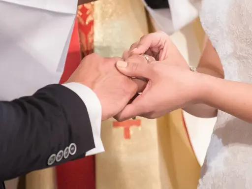 Dłoń panny młodej zakłada obrączkę na palec pana młodego. W tle widać księdza w szacie liturgicznej. To piękny ślub w innej parafii.