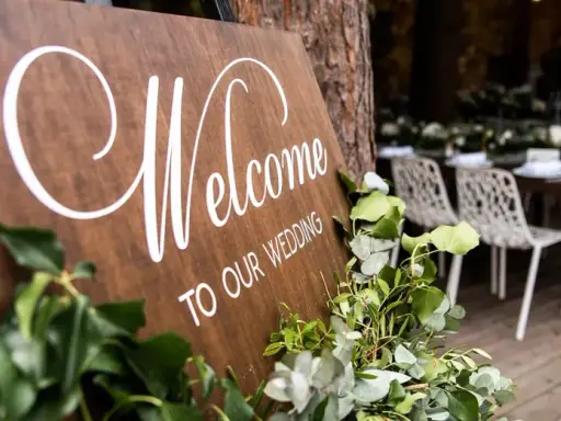 Drewniana tablica "Welcome to Our Wedding" ozdobiona zielenią. W tle widać przygotowane stoły i krzesła, a także obsługę. Opłata ZAIKS za muzykę na weselu.