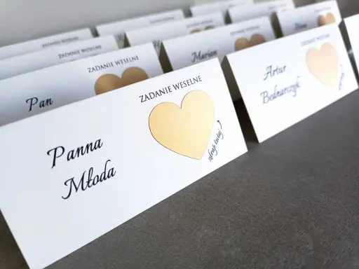 Białe karty z imionami gości i złotymi serduszkami kryją śmieszne zadania dla gości weselnych.