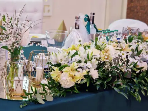 Elegancki pomysł na wesele: stół udekorowany bukietami białych i żółtych kwiatów, świecami w złotych lampionach i butelkami z napojami.