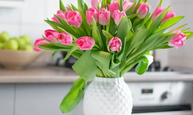 Różowe tulipany w białym wazonie, idealne na stół weselny. W tle miska z zielonymi jabłkami i kuchenny blat.