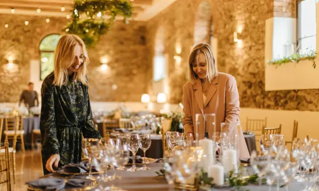 Dwie kobiety przygotowują salę na przyjęcie. Stół nakryty jest eleganckimi naczyniami, tworząc idealny scenariusz wesela.