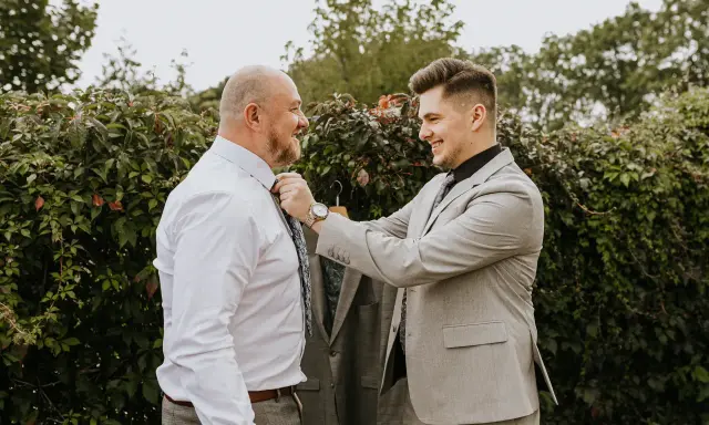 Przywitanie pana młodego w domu młodej. Młody mężczyzna w garniturze poprawia krawat starszemu, uśmiechniętemu panu.