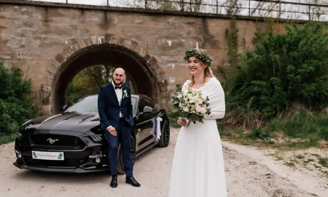 Para nietypowe sesje ślubne pozuje przy czarnym Mustangu pod kamiennym wiaduktem. Panna młoda w białej sukni i wianku z zieleni, pan młody w granatowym garniturze.