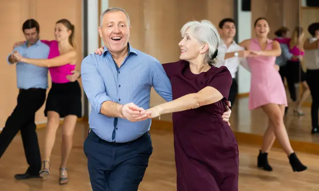 Uśmiechnięta para seniorów uczy się tańczyć w parze. W tle inne pary ćwiczą kroki.