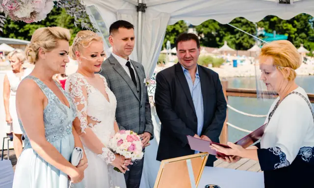 Urzędniczka w przyłbicy wręcza parze młodej dokumenty, symbolizujące ich licencję na ślub.